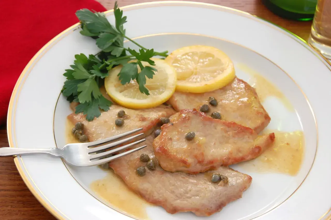 Piccata de veau, tendre escalope de veau sauce citronnée, câpres et persil frais, sur une assiette blanche bordée d'or.