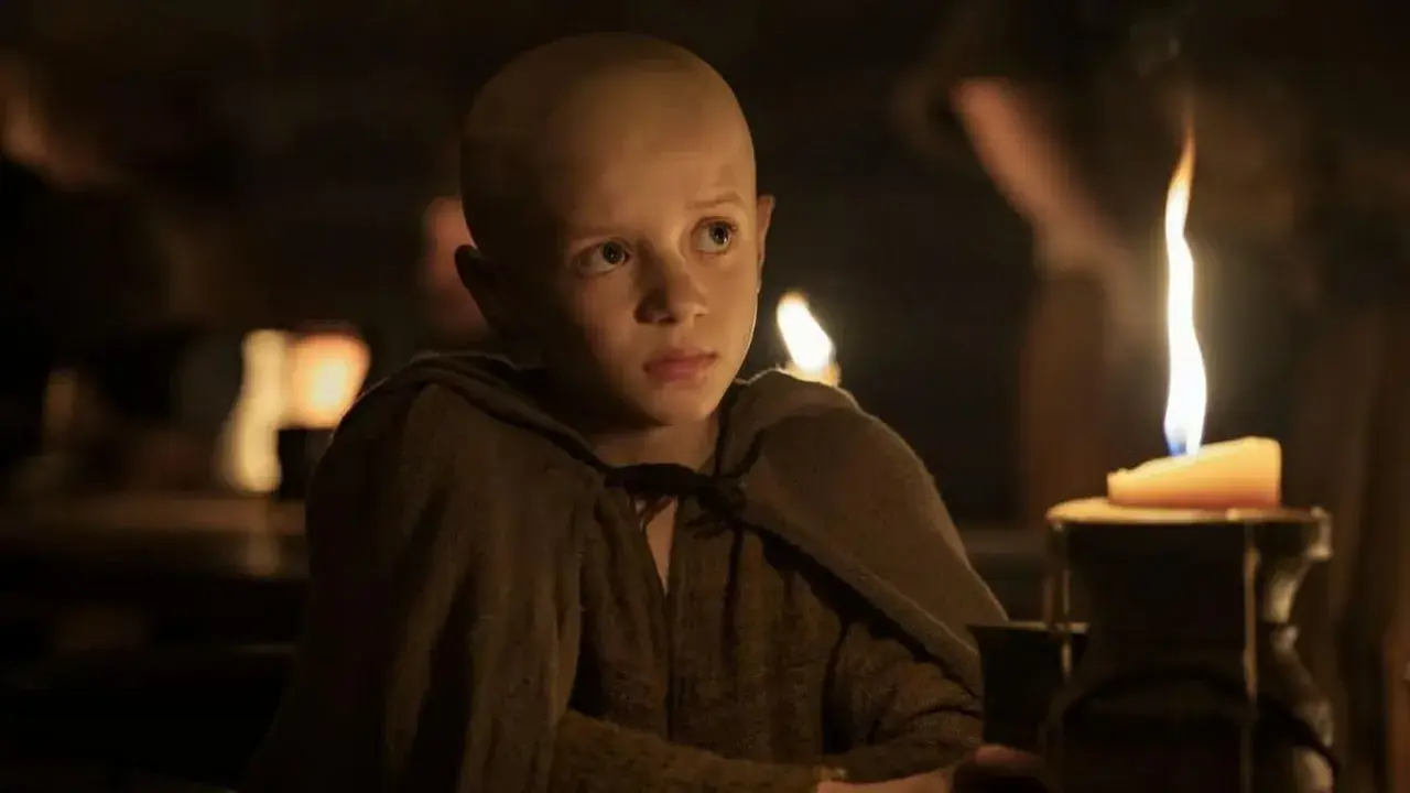 Un jeune garçon chauve, vêtu d'une tunique marron, regarde fixement une bougie allumée. Scène de "A Knight of the Seven Kingdoms Episode 3".