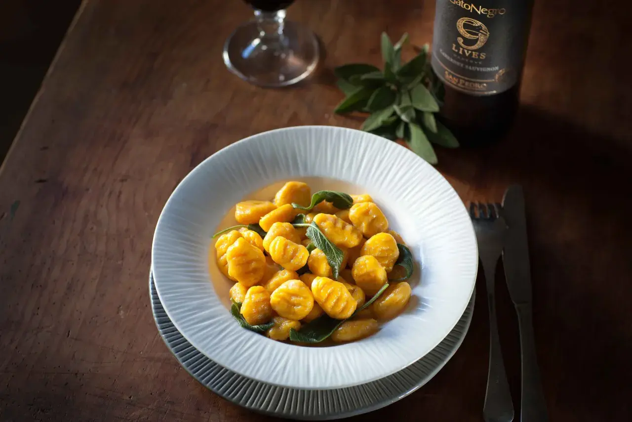 Gnocchis de potimarron aux feuilles de sauge, servis dans une assiette blanche sur une table en bois. Une bouteille de vin et un verre sont visibles en arrière-plan. Parfait pour une recette gnocchi potimarron.