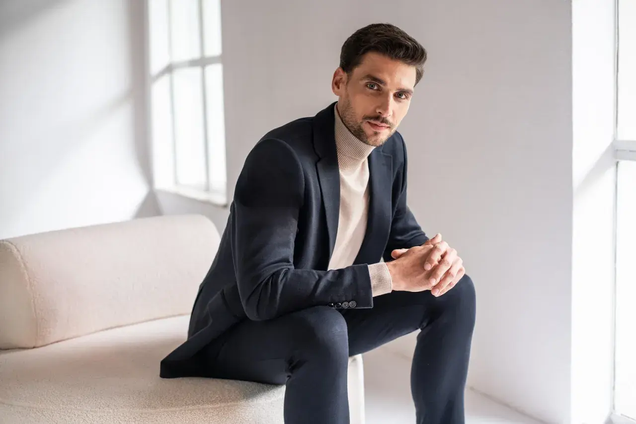 Mężczyzna w granatowym garniturze i beżowym golfie, prezentujący styl smart casual. Siedzi na kanapie, opierając dłonie na kolanach.
