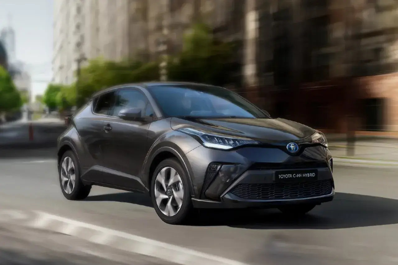 Ciemnoszary Toyota C-HR Hybrid w ruchu miejskim, z rozmytym tłem budynków i drzew.