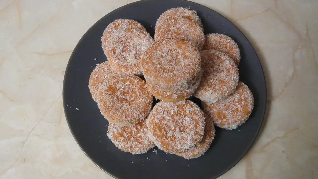 Montón de galletas de coco, doradas y cubiertas de coco rallado, apiladas en un plato oscuro.