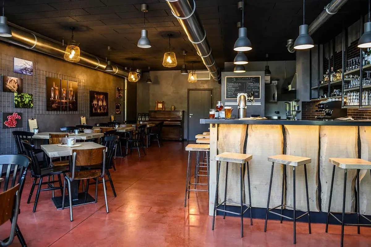 Interno di un locale con tavoli, sedie, un bancone in legno grezzo e sgabelli. Sul bancone due birre, pronte per essere gustate dal baldo birraio.