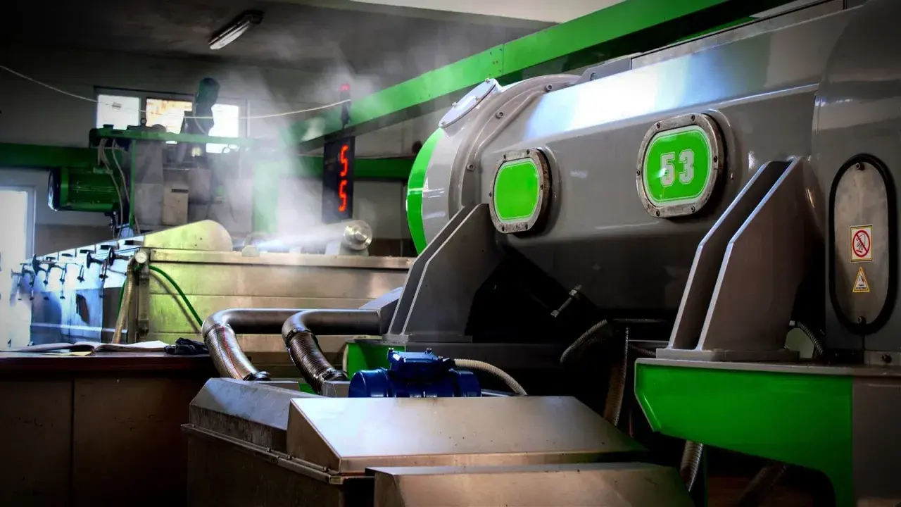 Macchinari industriali in una fabbrica, con tubi e vapore. Si intravede un cartello con "53", forse legato alla produzione di olio di sansa cos'è.