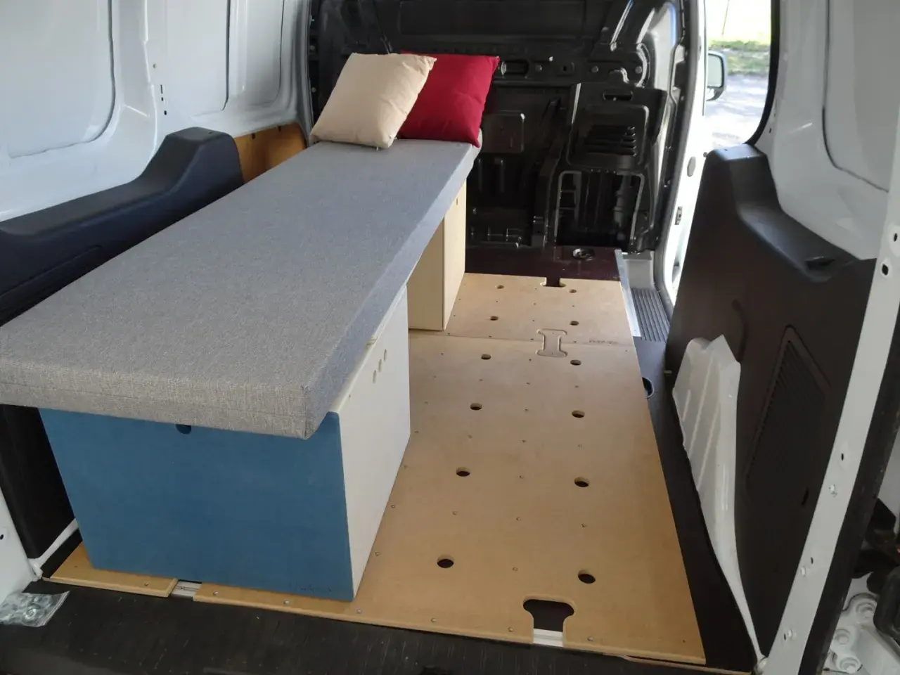 Aménagement intérieur d'un petit fourgon : banquette grise avec coussins, caisson de rangement bleu et blanc, plancher en bois. Prêt pour l'aventure !