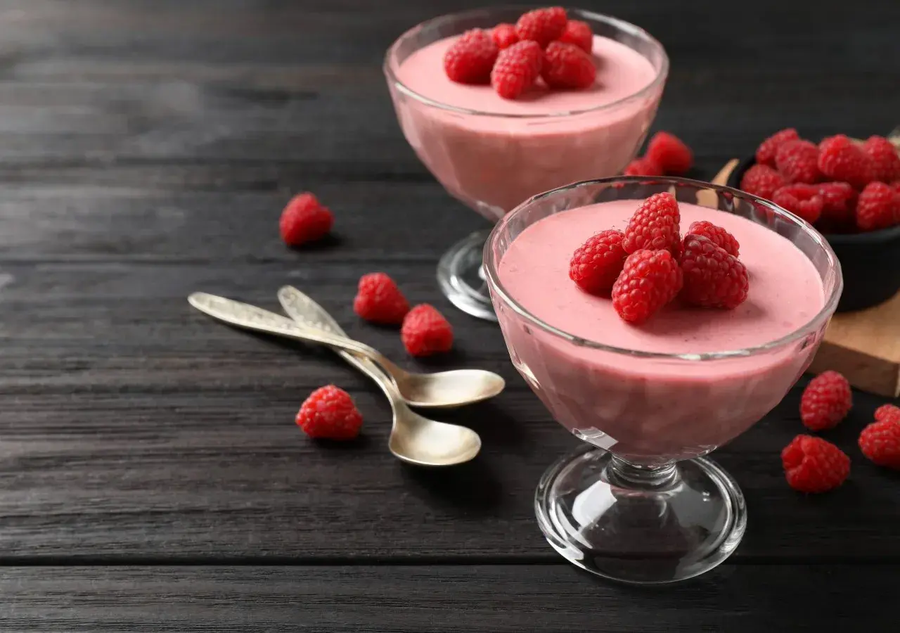 Två glas med hallonmousse, toppade med färska hallon. Två skedar ligger bredvid på ett mörkt träbord.