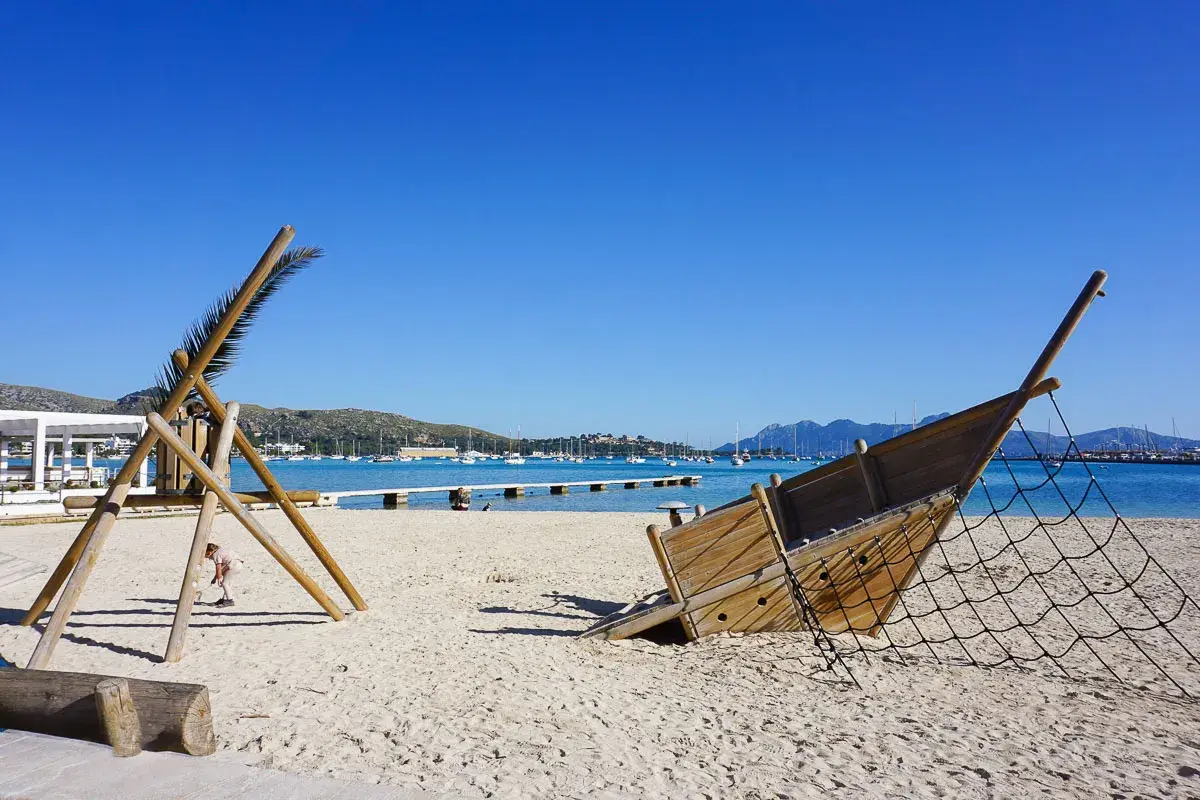 Spielplatz am Strand im mallorcas norden. Ein hölzernes Schiffswrack und eine Kletterstruktur stehen im Sand, Boote liegen im türkisfarbenen Wasser.