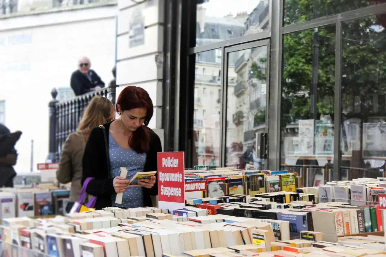 Une jeune femme choisit un livre dans une librairie en plein air, devant une vitrine reflétant des bâtiments parisiens. Une sélection de livres, dont des romans policiers et de suspense, est exposée, rappelant une maison d'édition jeunesse.