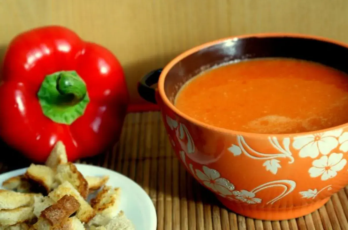 Aksamitny zupa krem z papryki, podana z chrupiącymi grzankami. Obok leży świeża, czerwona papryka.