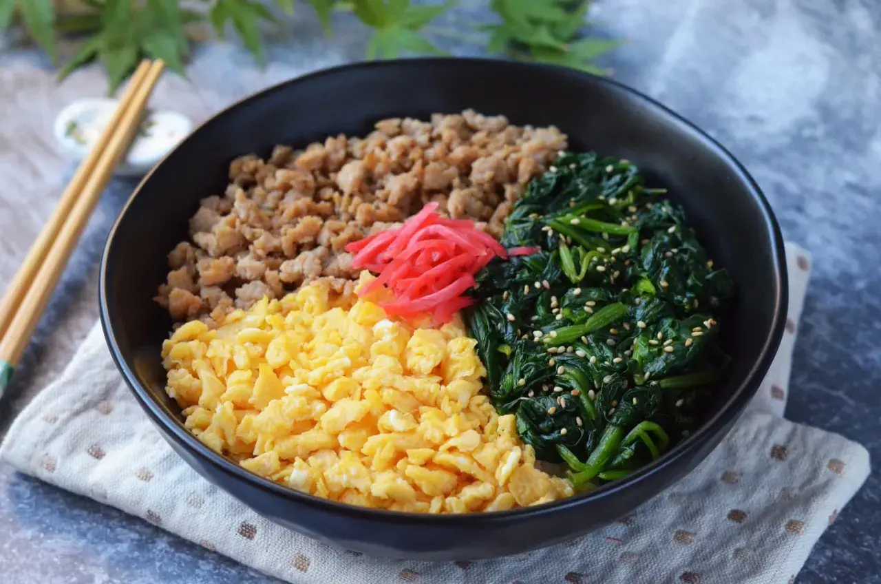 A colorful sanshoku don bowl with scrambled eggs, seasoned ground meat, and spinach, topped with pickled ginger.