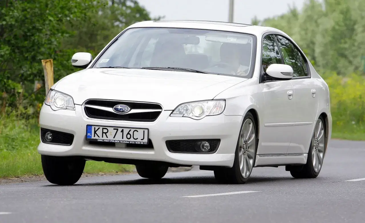 Białe Subaru Legacy z polskimi tablicami KR 716CJ jedzie po drodze wśród zieleni.