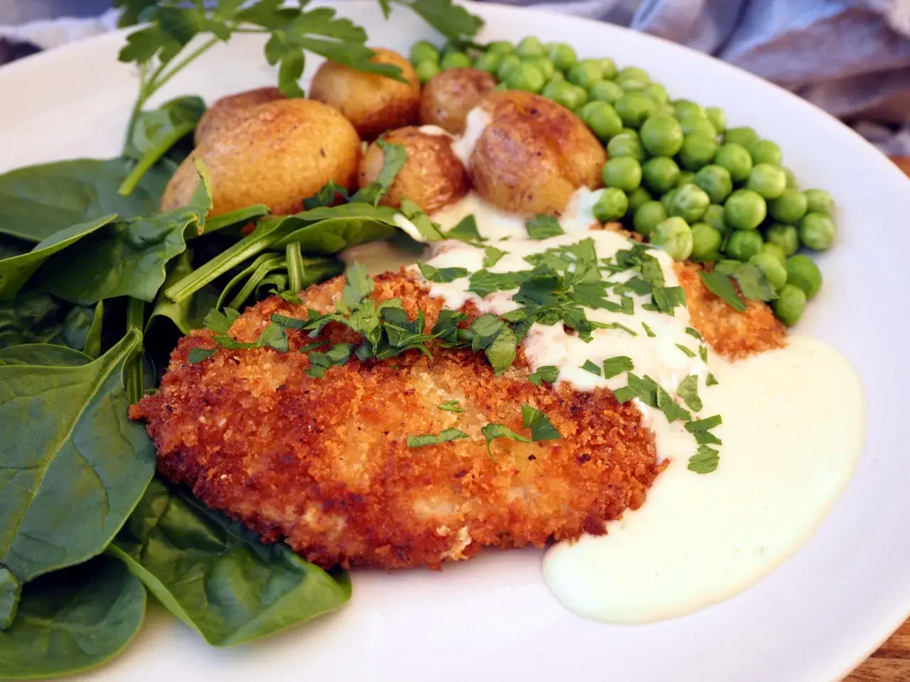 Krispig schnitzel med krämig sås till, serverad med småpotatis, gröna ärtor och färsk spenat.