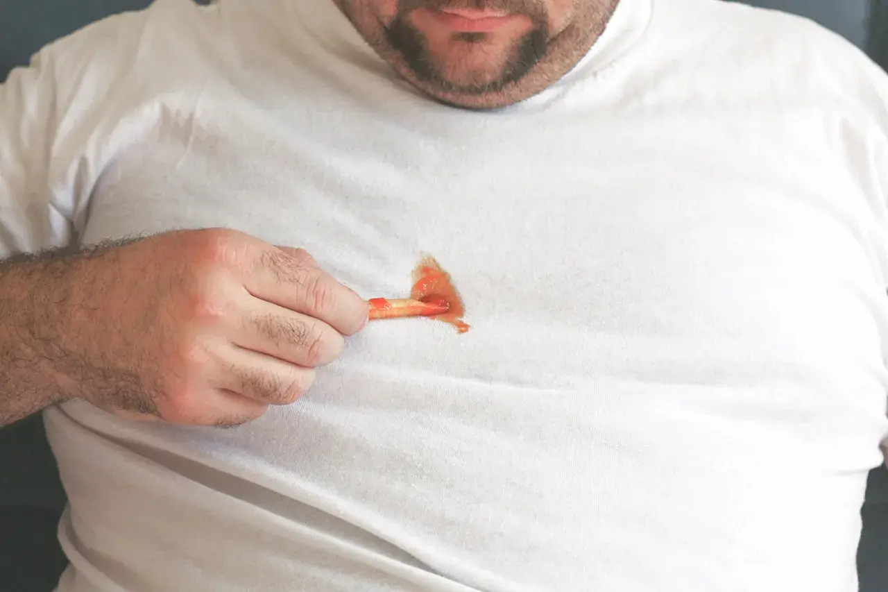 Ein Mann zeigt auf einen Ketchupfleck auf seinem weißen T-Shirt. Solche Fettflecken auf Kleidung sind ärgerlich.