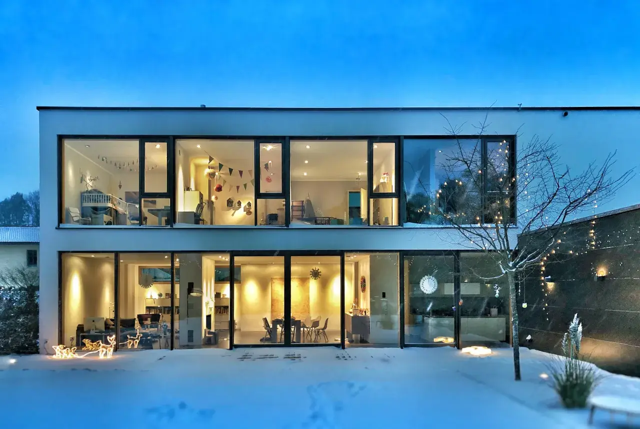 Casa moderna con grandes ventanales iluminados, mostrando interiores acogedores. La nieve cubre el exterior, y luces navideñas decoran un árbol. ¡2500 lumens cuanto ilumina!