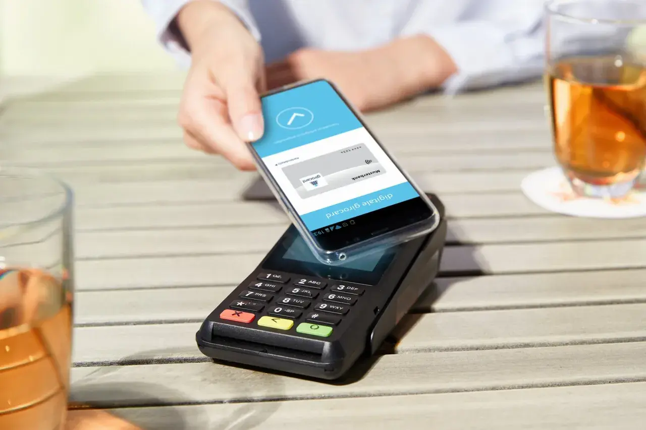 Mit handy bezahlen: Eine Hand hält ein Smartphone über ein Kassenterminal, um kontaktlos zu bezahlen.