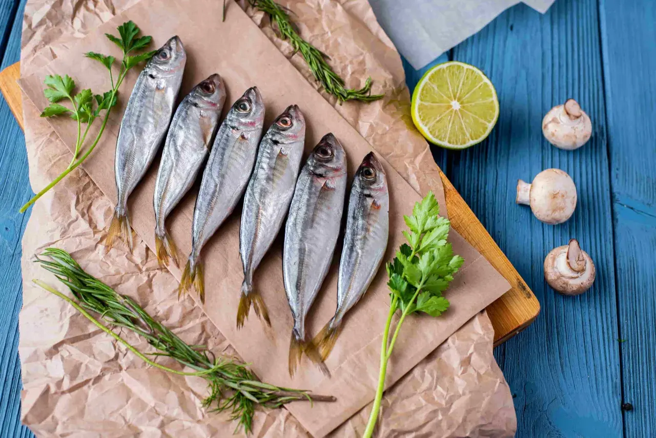 Seis jureles pescado frescos, listos para cocinar, acompañados de perejil, romero, limón y champiñones sobre papel de estraza y tabla de madera.