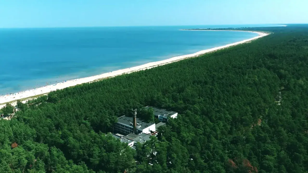 Widok z lotu ptaka na hotel otoczony lasem sosnowym, z plażą i morzem w tle.