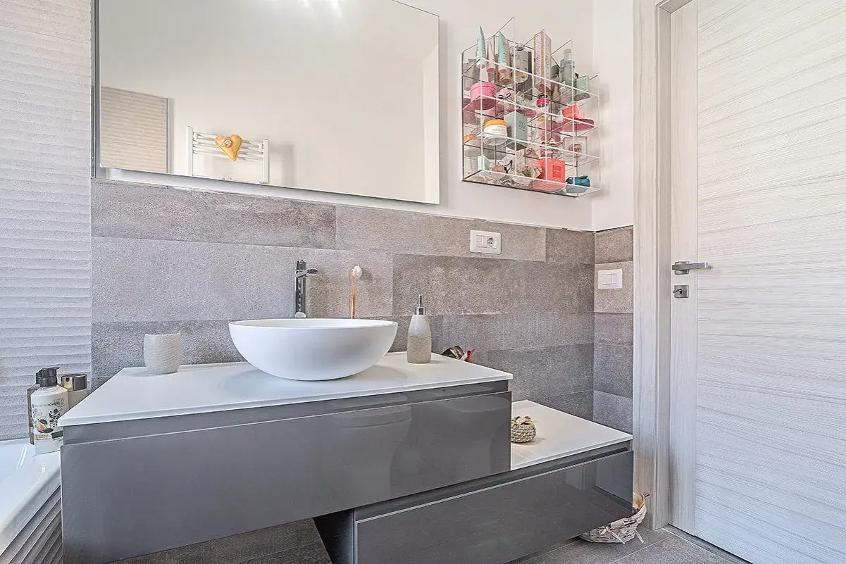 Bagno moderno grigio e legno con lavabo bianco, specchio e porta in legno chiaro. Organizzatore trasparente pieno di cosmetici.