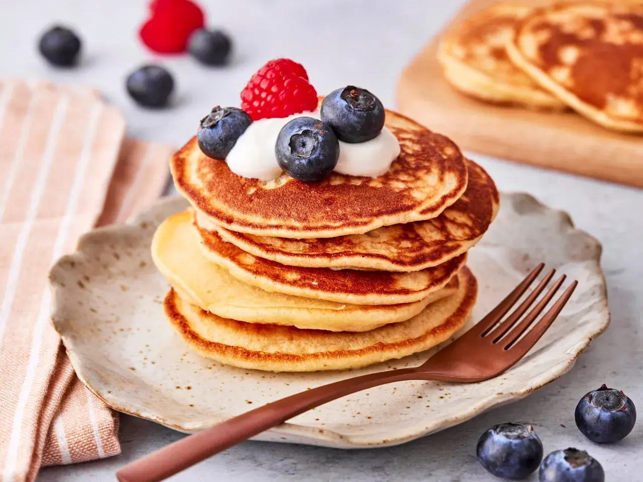 Ein Stapel goldbrauner Pancakes mit Joghurt, Blaubeeren und einer Himbeere obendrauf. Ein köstlicher pancake mit joghurt für ein perfektes Frühstück.