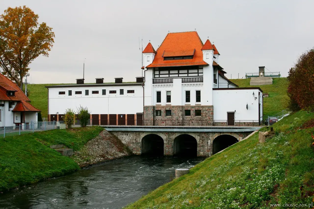 Elektrownia wodna Straszyn z charakterystycznym czerwonym dachem i kamienną podstawą, nad rwącą rzeką.