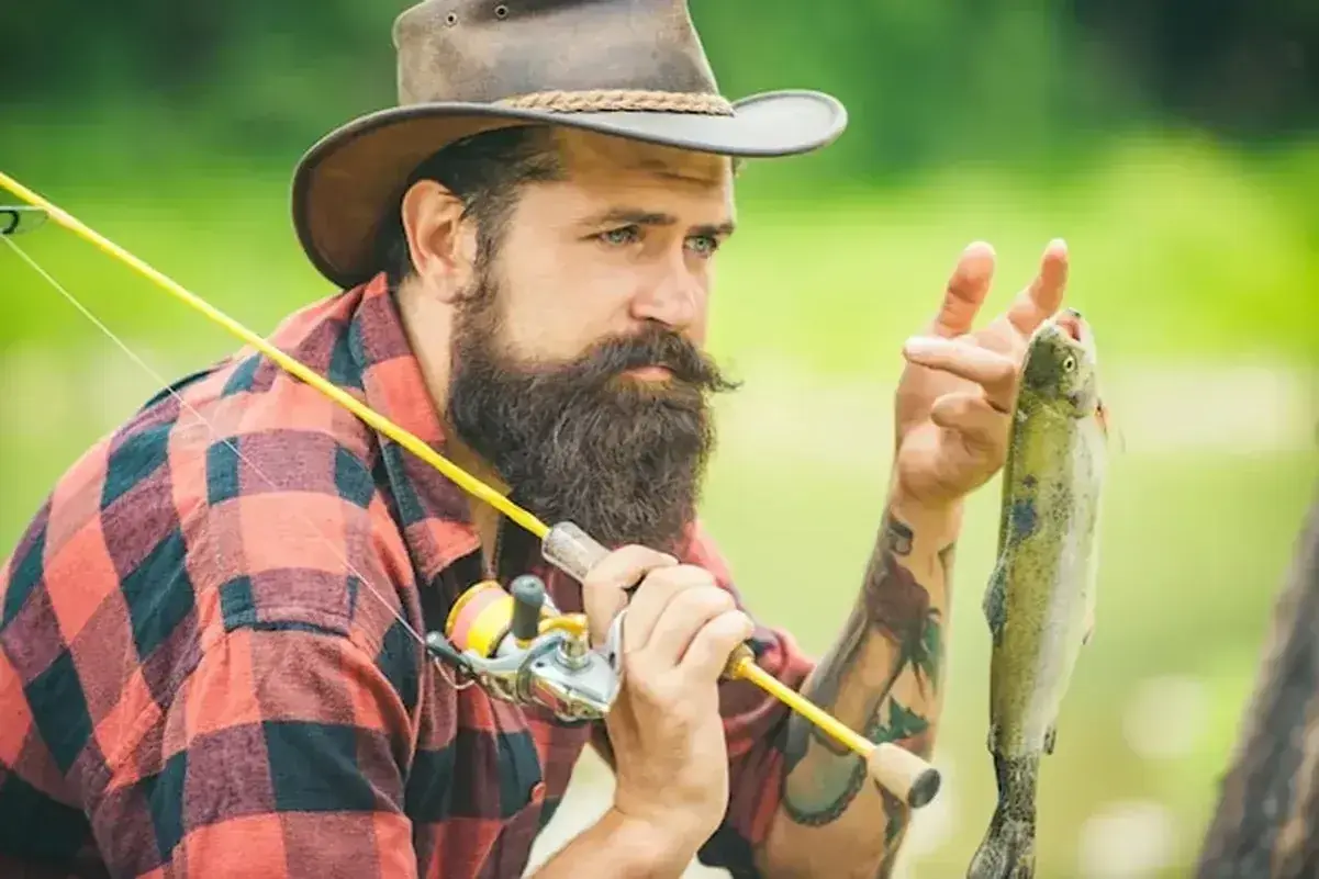 Brodaty wędkarz w kapeluszu trzyma świeżo złowionego pstrąga. Idealna pogoda wędkarska.