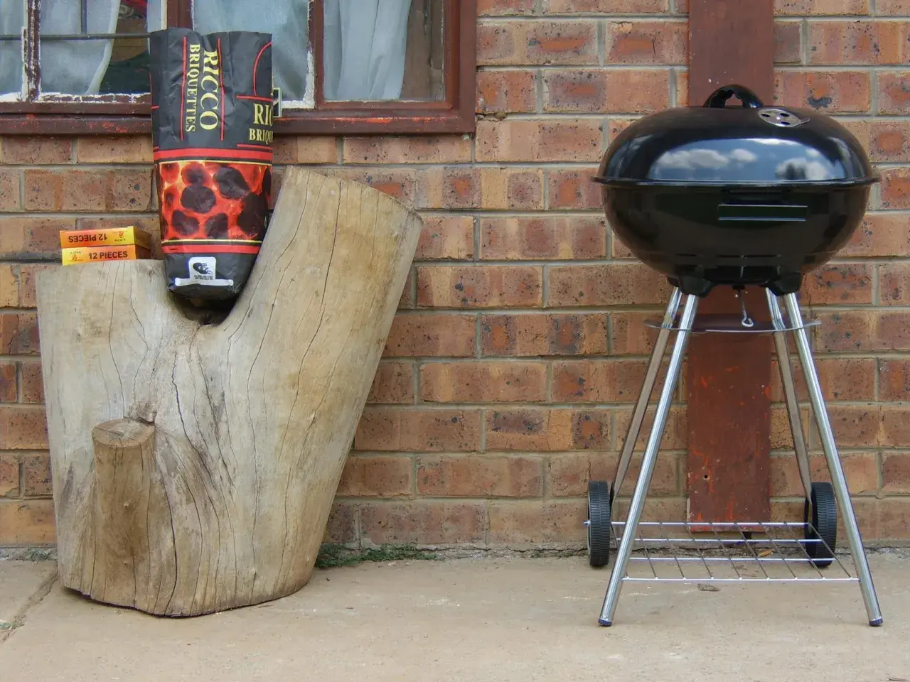 Grill stoi obok pnia, a torba z węglem drzewnym wisi na pniu. Odległość grilla od granicy działki jest bezpieczna.