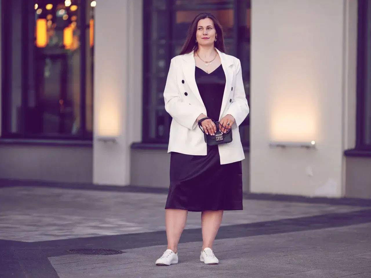Eine Frau in einem schwarzen Kleid und weißem Blazer, die zeigt, was man zum Musical anzieht. Sie trägt weiße Sneaker und eine kleine Handtasche.
