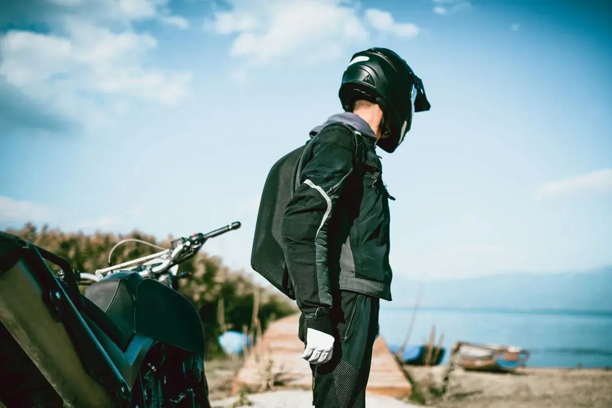 Motociclista con casco negro, chaqueta y guantes blancos, junto a su moto. El tejido de poliéster 600d vs Cordura es ideal para su aventura.