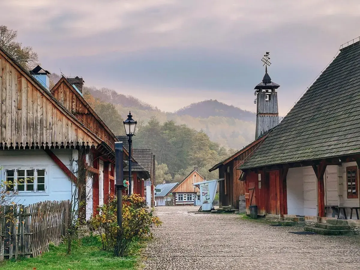 Sanok skansen: urokliwa, brukowana uliczka z drewnianymi domami i wieżą z wiatrowskazem, otoczona mglistymi wzgórzami.