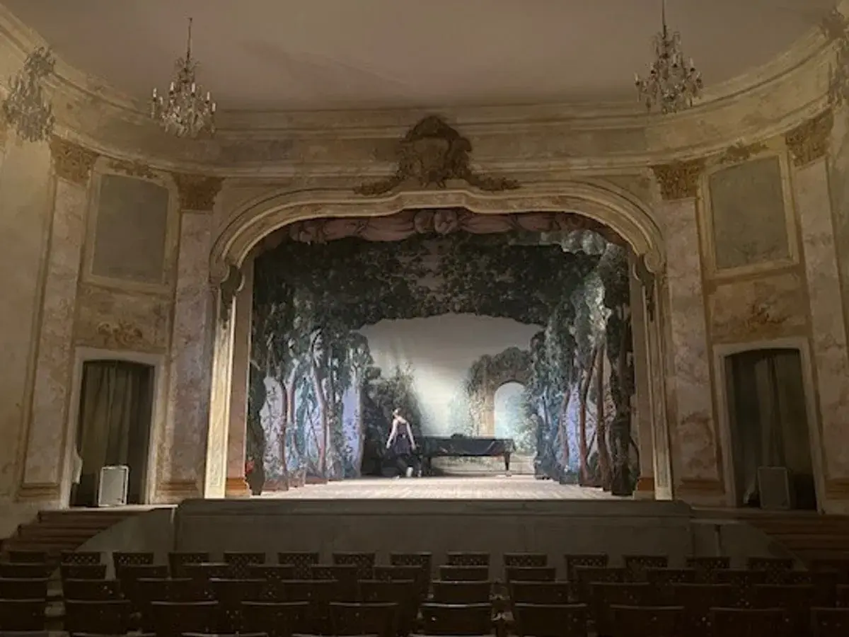 En ensam person vid ett piano på en scen med en skogsmålning, i den vackra teatersalongen på **Confidencen Ulriksdal**.