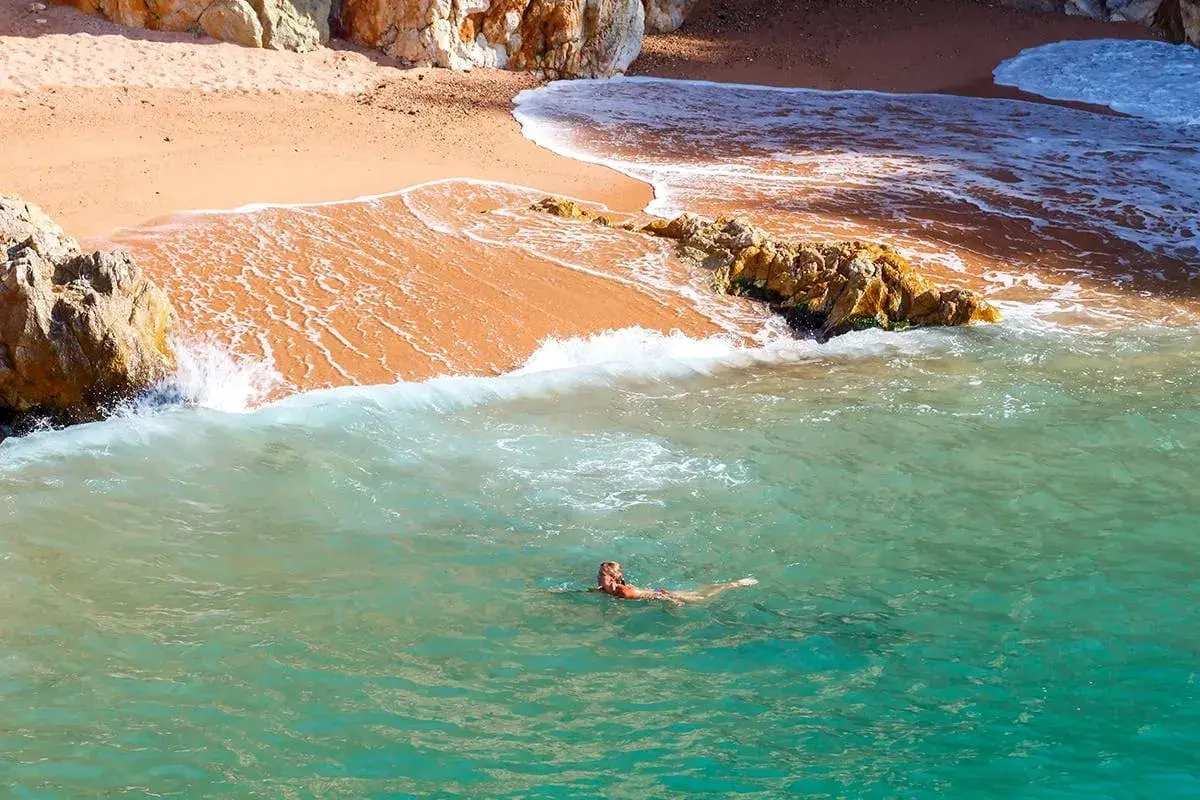 Eine Frau schwimmt im türkisfarbenen Wasser vor einem goldenen costa brava strand, umgeben von Felsen.