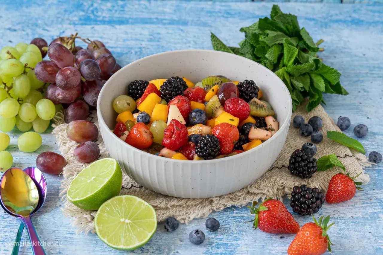 Ein bunter, sommerlicher Obstsalat mit Beeren, Trauben und Mango, garniert mit Minze und Limettenspalten.