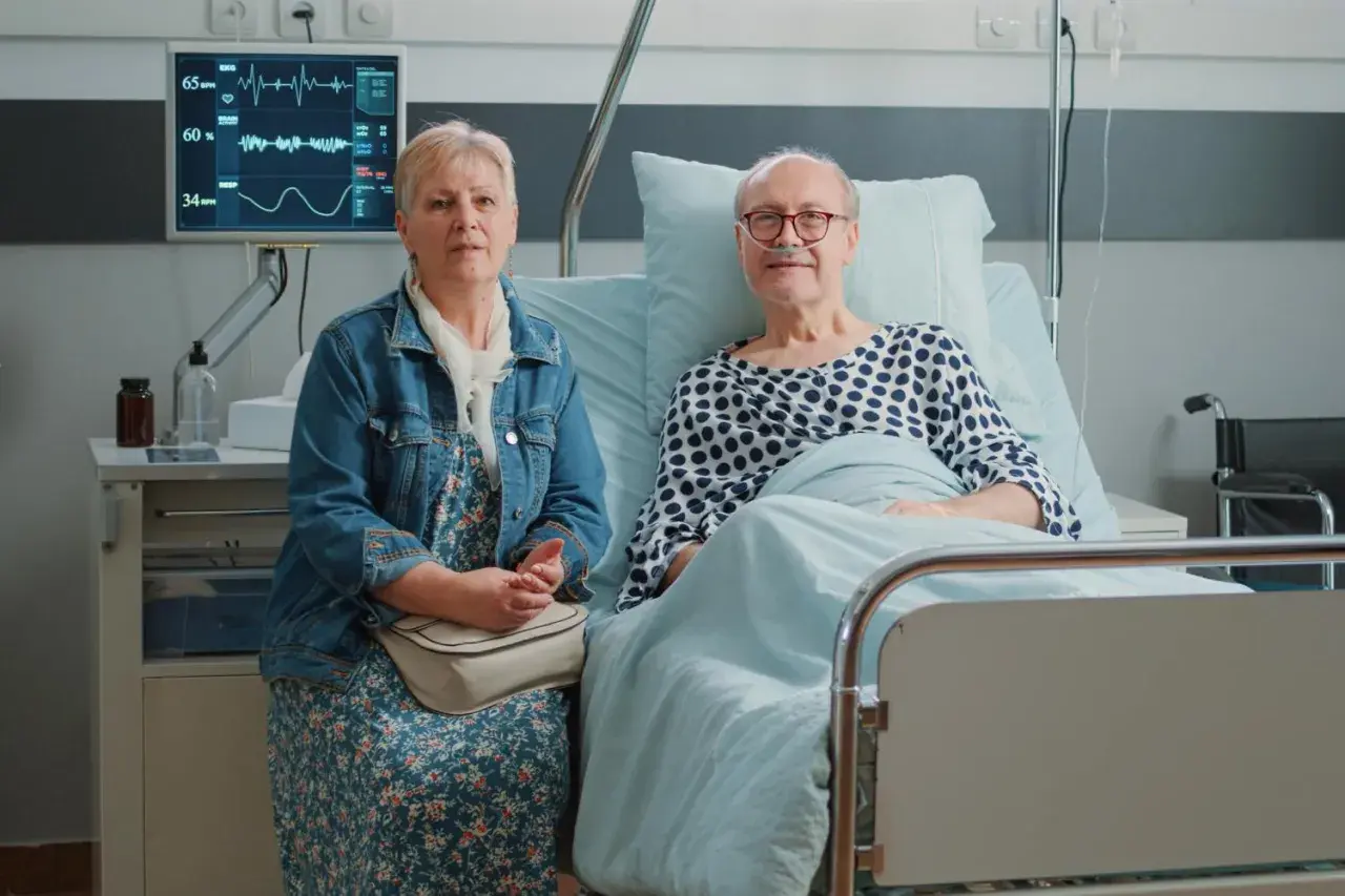 Starsza kobieta siedzi obok starszego mężczyzny w łóżku szpitalnym, na monitorze widoczne są parametry życiowe.