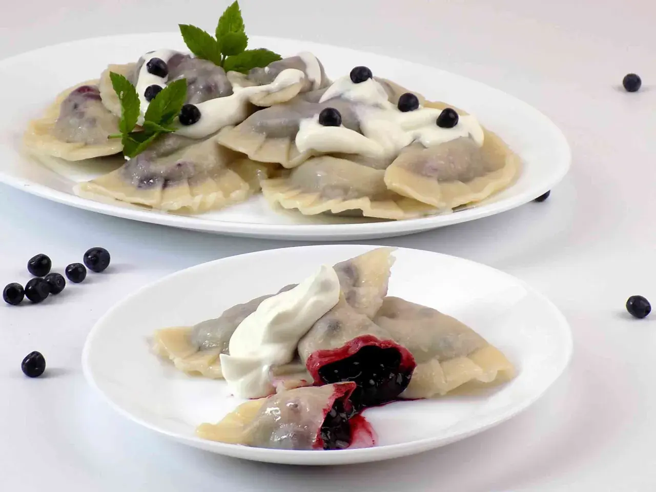 Pyszne pierogi z jagodami, polane śmietaną i ozdobione miętą. Na talerzu widać też kilka świeżych jagód.
