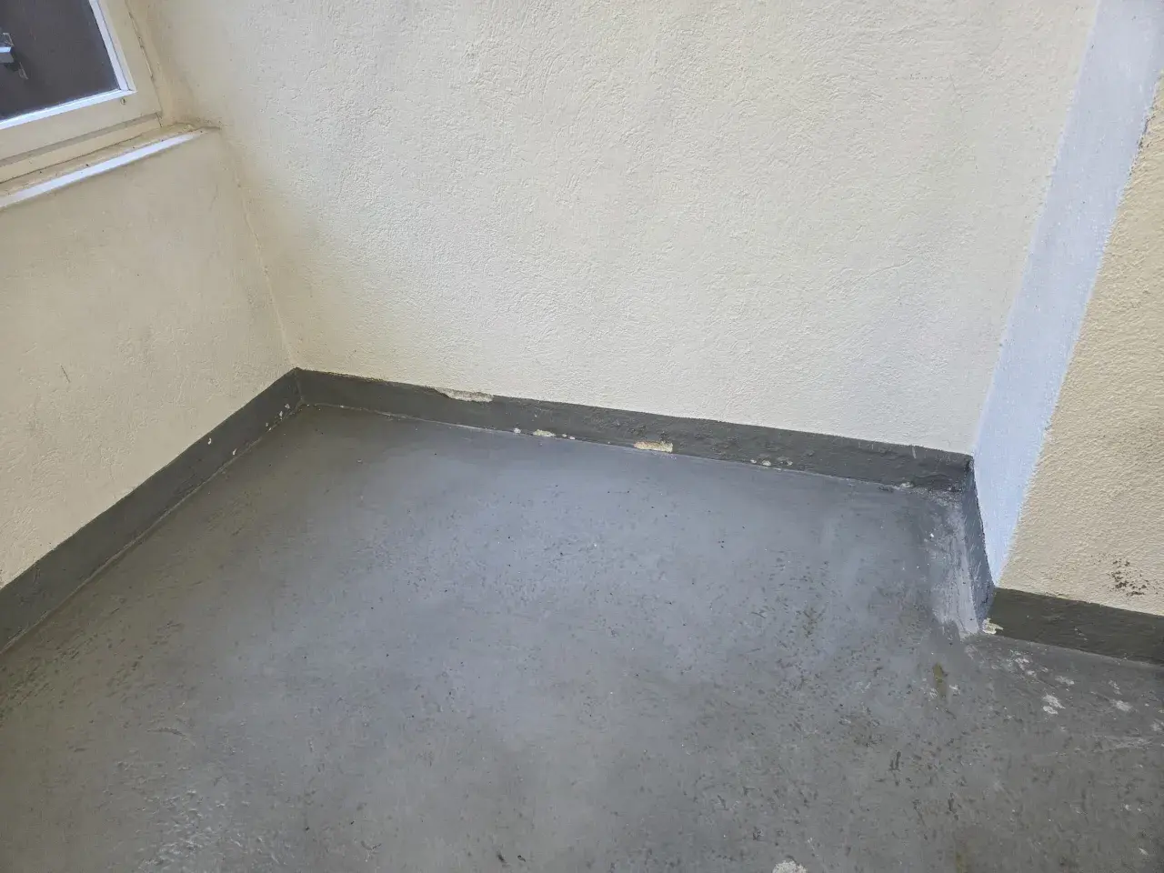 Ecke eines Raumes mit grauem Boden und weißer Wand. Am Sockel ist ein dunkler Streifen zu sehen, auf dem sich schimmel zieht von unten nach oben.