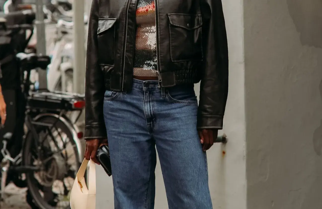 Personne portant une veste en cuir noire, un haut à paillettes et un jean bleu, tenant un sac beige.