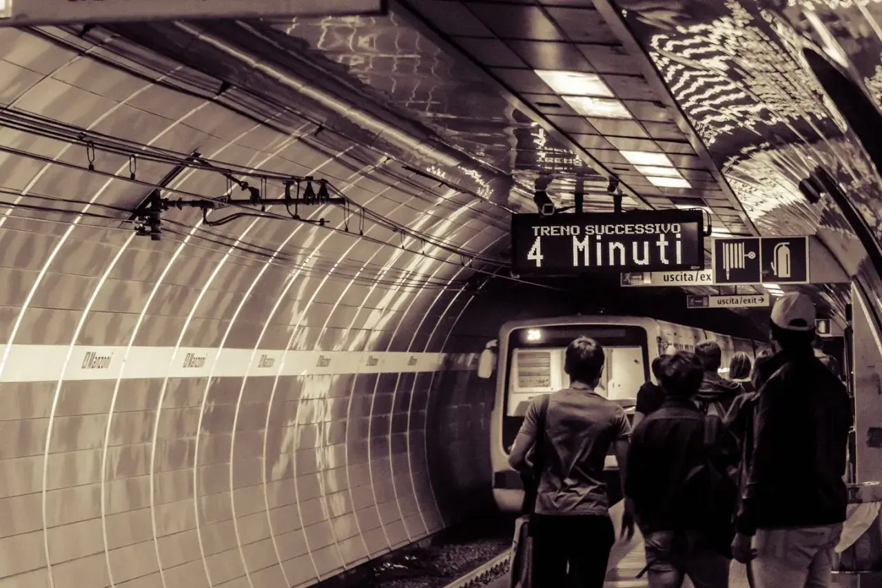 Persone attendono il treno sulla metro linea b. Un cartello indica "Treno successivo: 4 minuti".