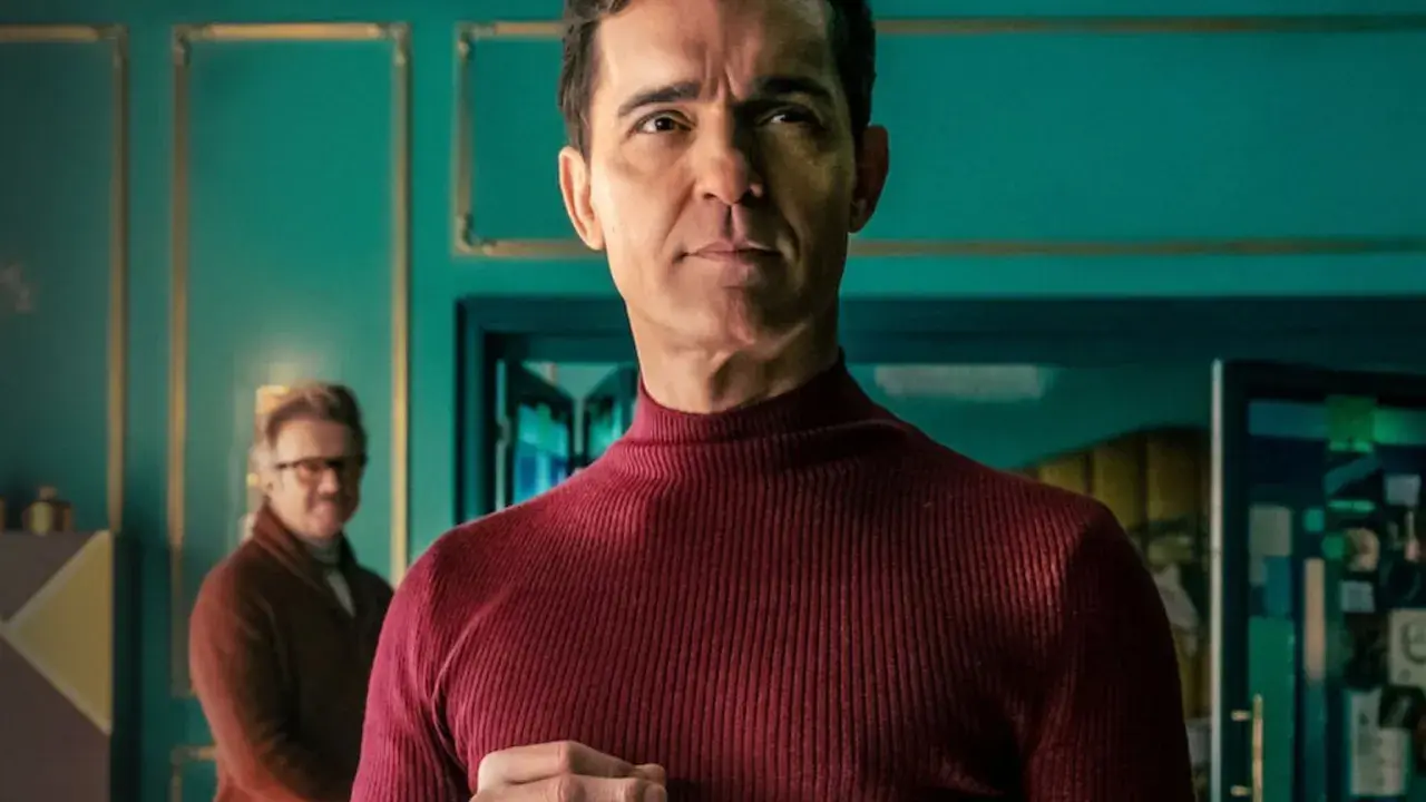 Berlin z "La Casa de Papel" w bordowym golfie, zamyślony. W tle inny mężczyzna.