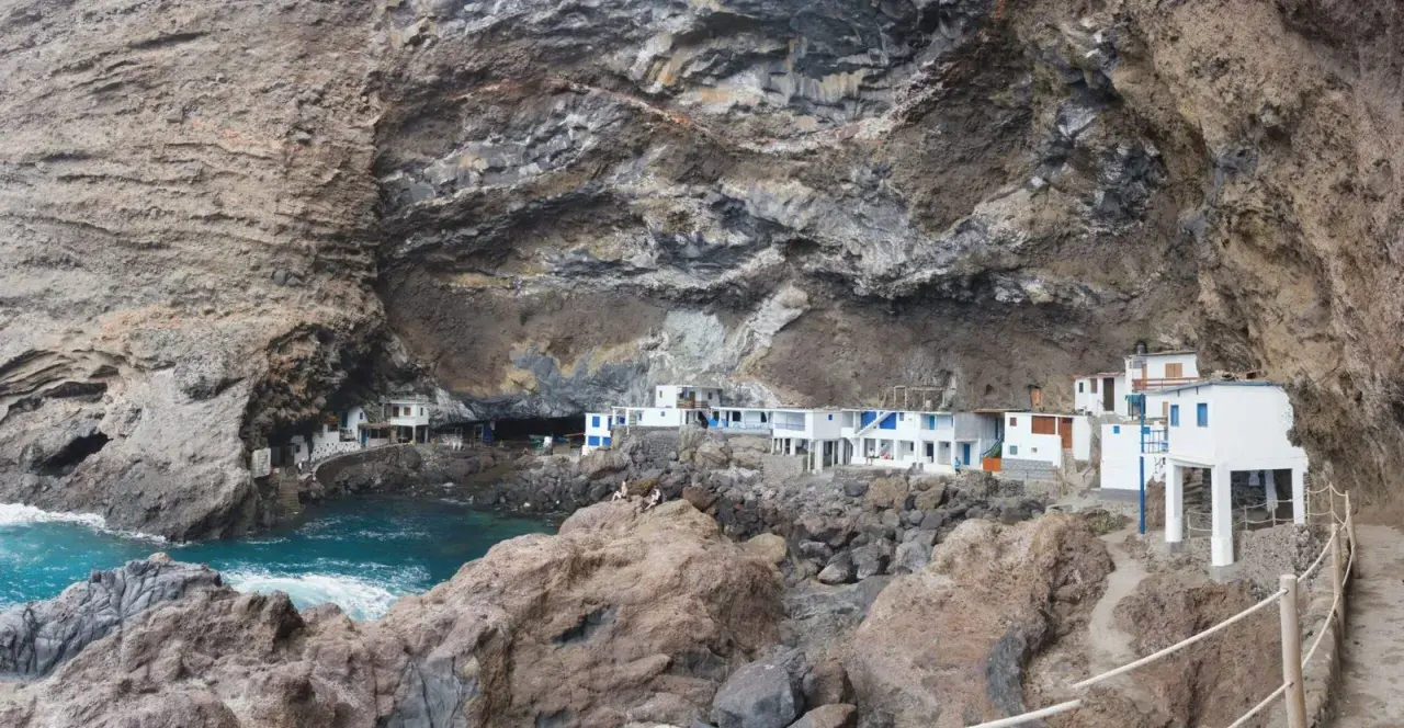 Casas blancas con detalles azules se asoman desde una cueva en los poris de Candelaria, La Palma. El mar turquesa rompe contra las rocas.