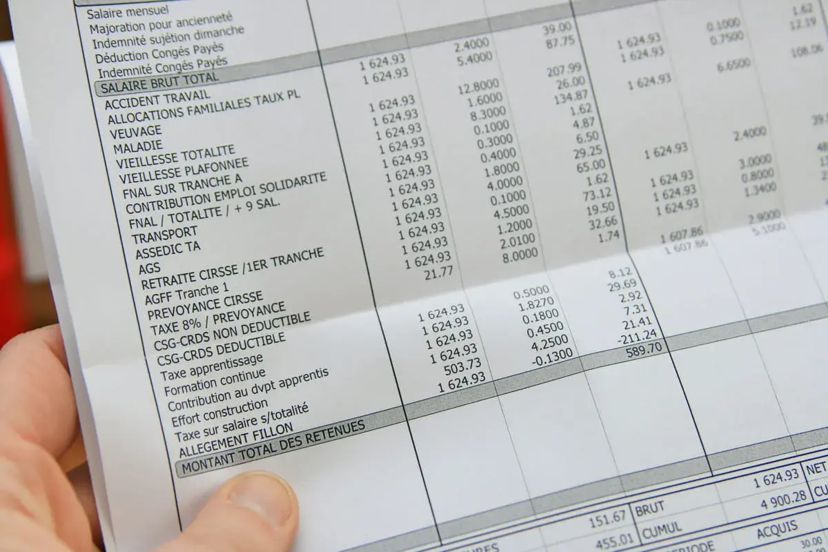 Une main tient une fiche de paie perdue, détaillant les salaires et les retenues.