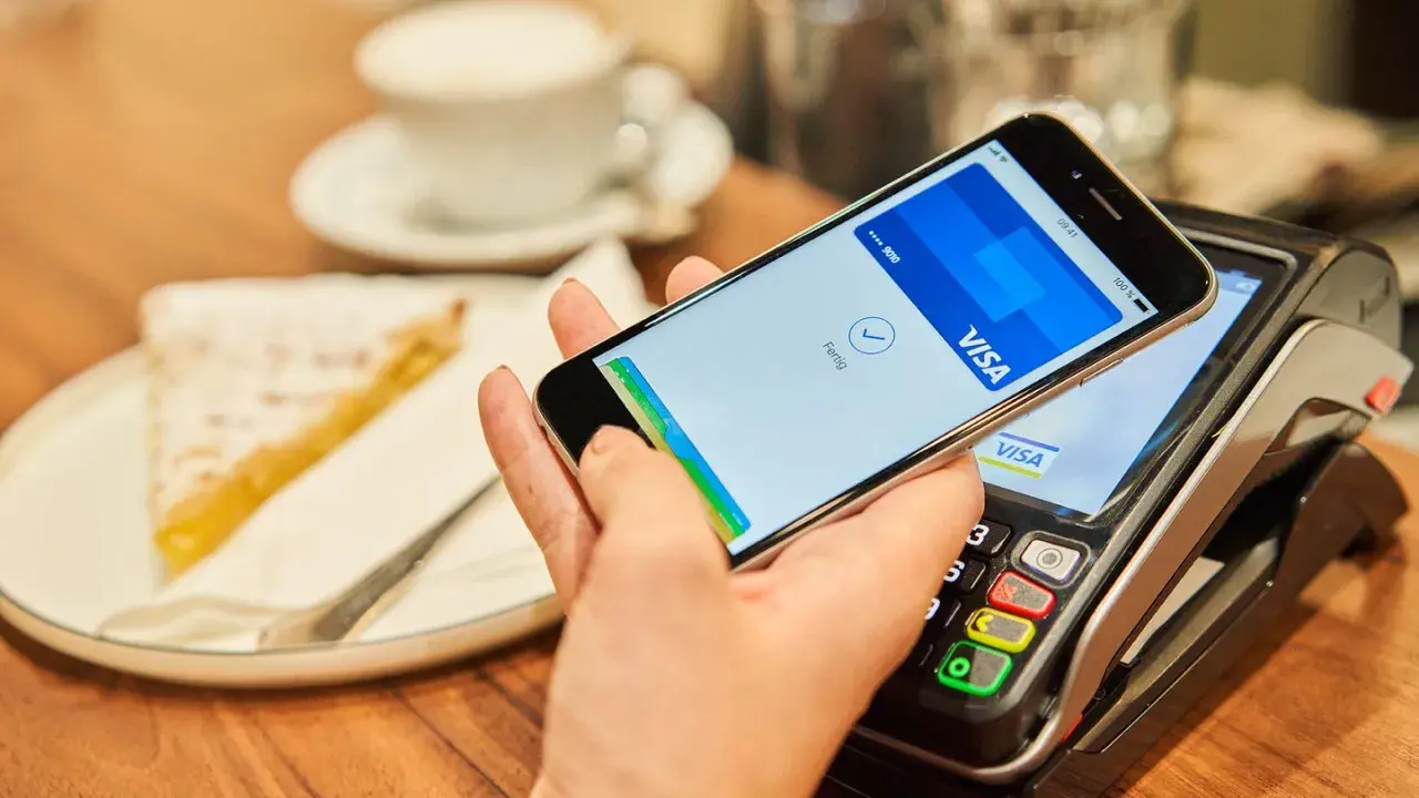Bezahlvorgang mit Apple Pay: Ein Smartphone hält über einem Kartenlesegerät, daneben ein Stück Kuchen.