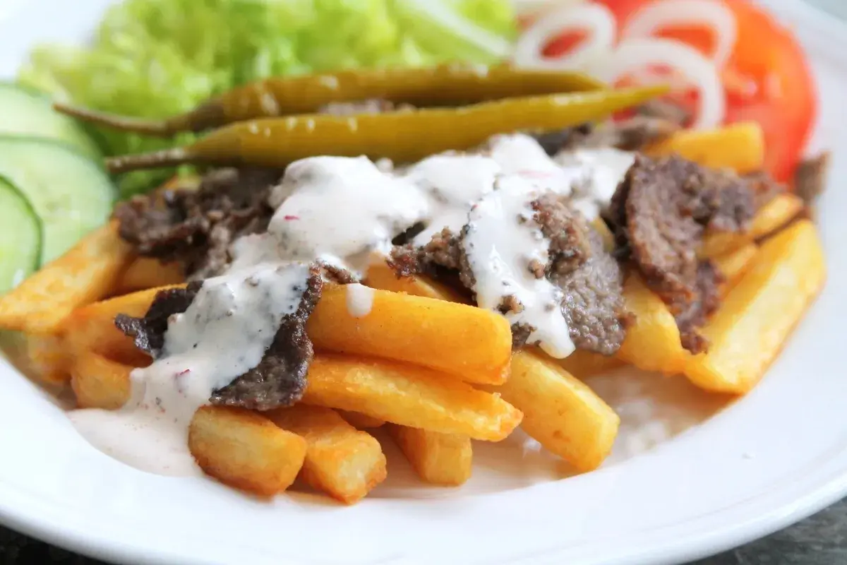 En tallrik med pommes frites, kebabkött och vit sås. Tillbehör som sallad, gurka, tomat, lök och jalapeños. Ett gott kebab recept!