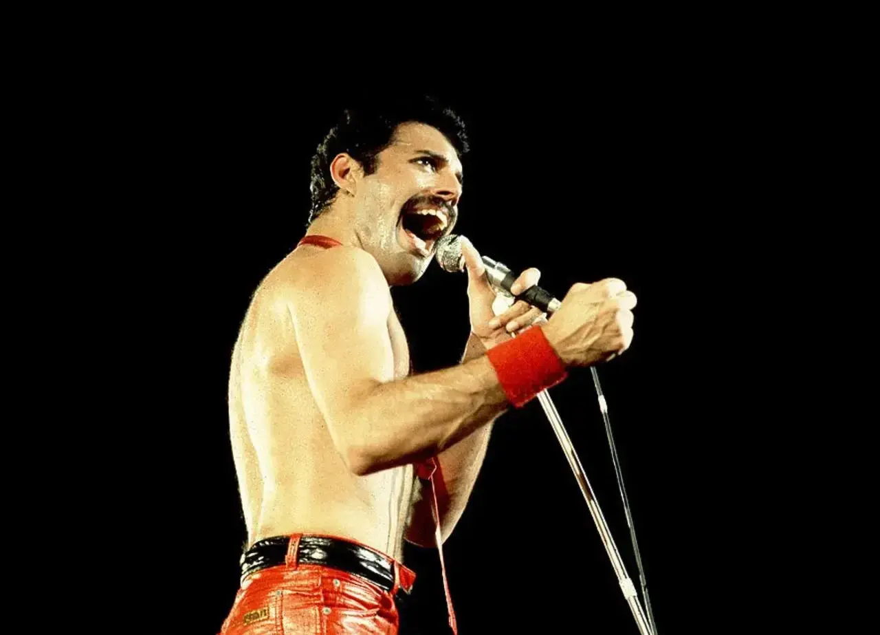 Freddie Mercury w czerwonych spodniach i opasce na nadgarstku śpiewa do mikrofonu. Nawet jako dorosły, jego energia przypominała dzikie dziecięce zabawy.