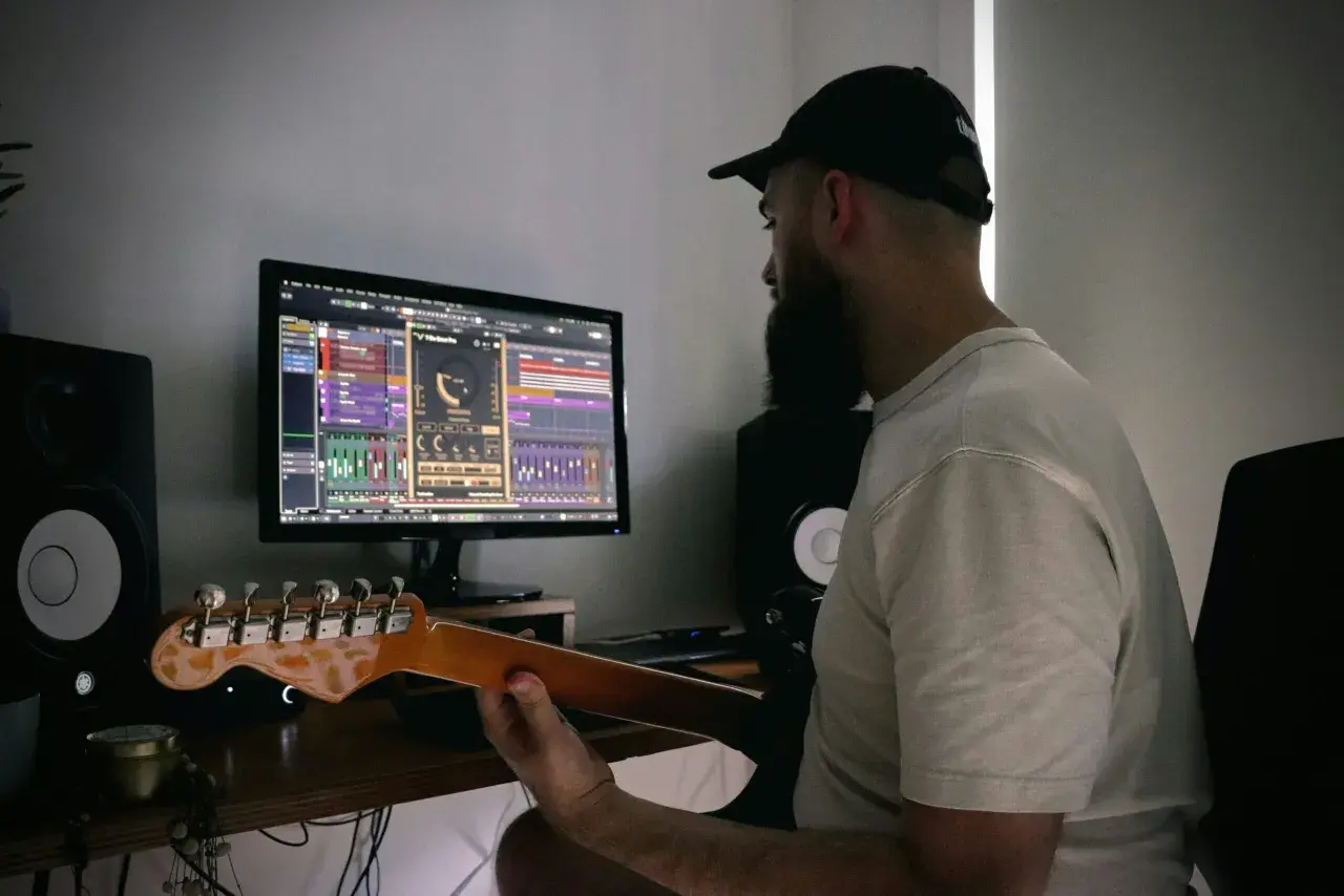 Mężczyzna z brodą i czapką z daszkiem uczy się, jak podłączyć gitarę do PC, pracując nad muzyką na komputerze.