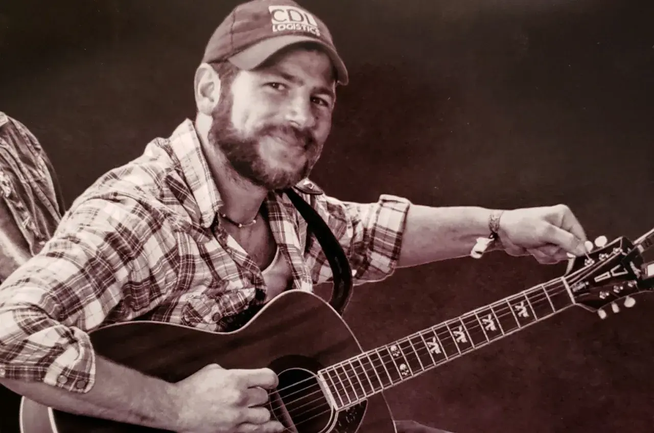 Kyle Shearer, a bearded man in a plaid shirt and cap, strums his acoustic guitar, ready to play a tune.