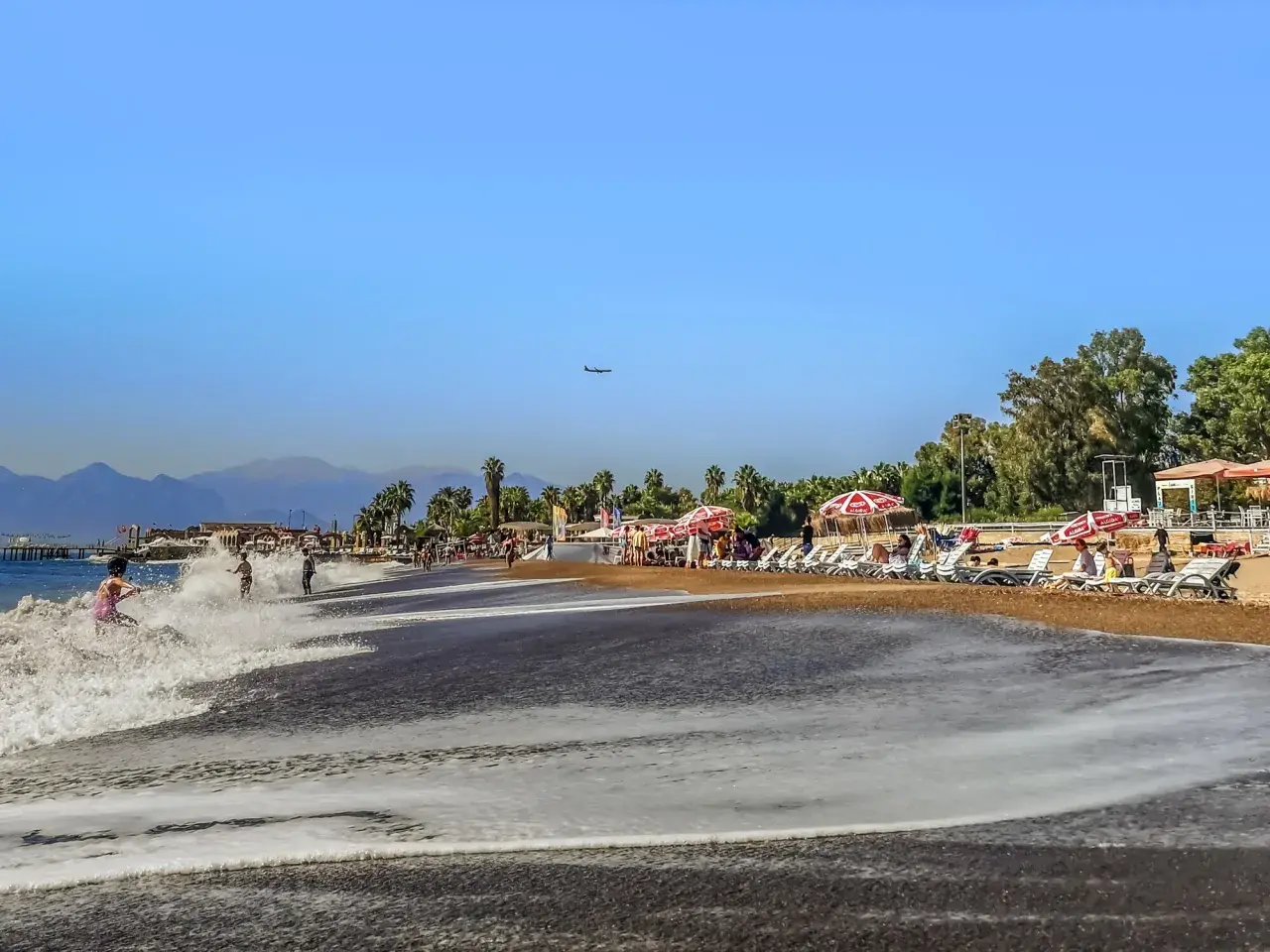 Ein sonniger Antalya Strand mit sanften Wellen, Liegestühlen und Palmen. Ein Flugzeug zieht seine Bahn am blauen Himmel.