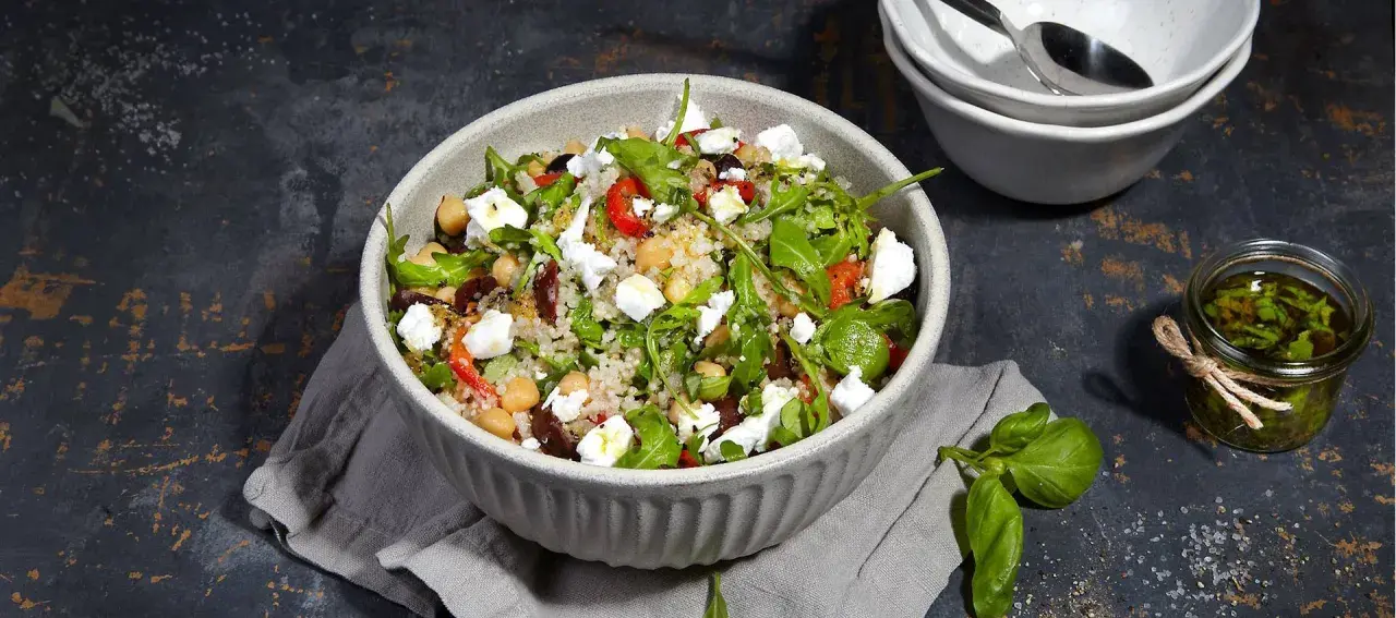 Bunter Quinoa Salat mit Kichererbsen, Feta, Oliven und Rucola, serviert in einer Schüssel. Daneben ein Glas mit Kräuter Dressing.
