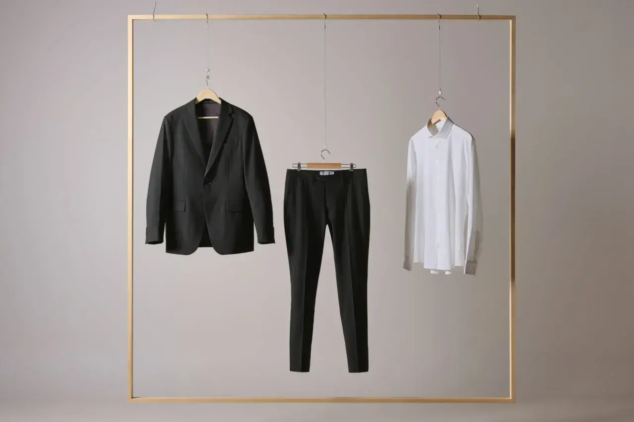 Ein dunkler Anzug, eine schwarze Hose und ein weißes Hemd hängen an einem Holzrahmen. Die Hose zeigt, wie lang eine Anzughose sein muss.