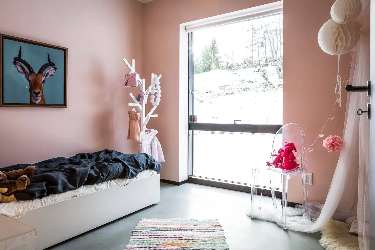 Ein gemütliches Kinderzimmer mit einer rosa Wand, einem Bett, Spielzeug und einem großen Fenster mit Blick auf eine verschneite Landschaft.