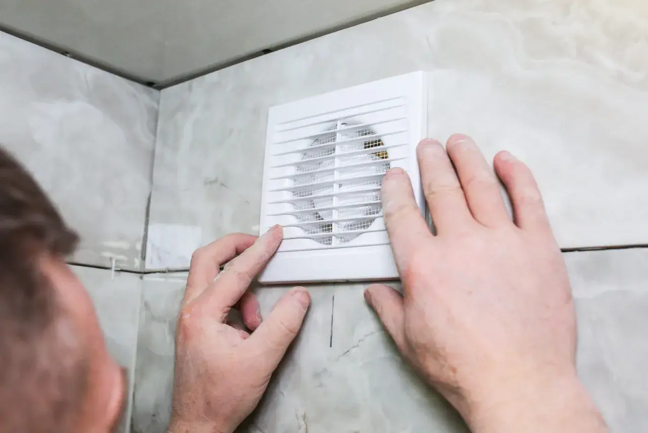Installation d'un ventilateur pour l'aération de la salle de bain. Des mains ajustent l'appareil blanc sur un mur carrelé.